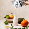 Person pouring a olive oil from a pouch into a white ceramic olive oil bottle with fruits and vegetables on a table.