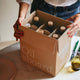 A person holds an open cardboard box labeled 'Oil aboard' containing several dark glass bottles with cork stoppers, set on a light marble countertop in a kitchen-like setting.