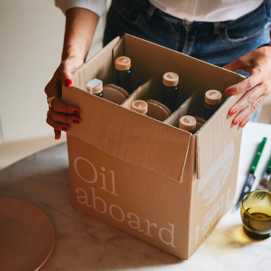 A person holds an open cardboard box labeled 'Oil aboard' containing several dark glass bottles with cork stoppers, set on a light marble countertop in a kitchen-like setting.