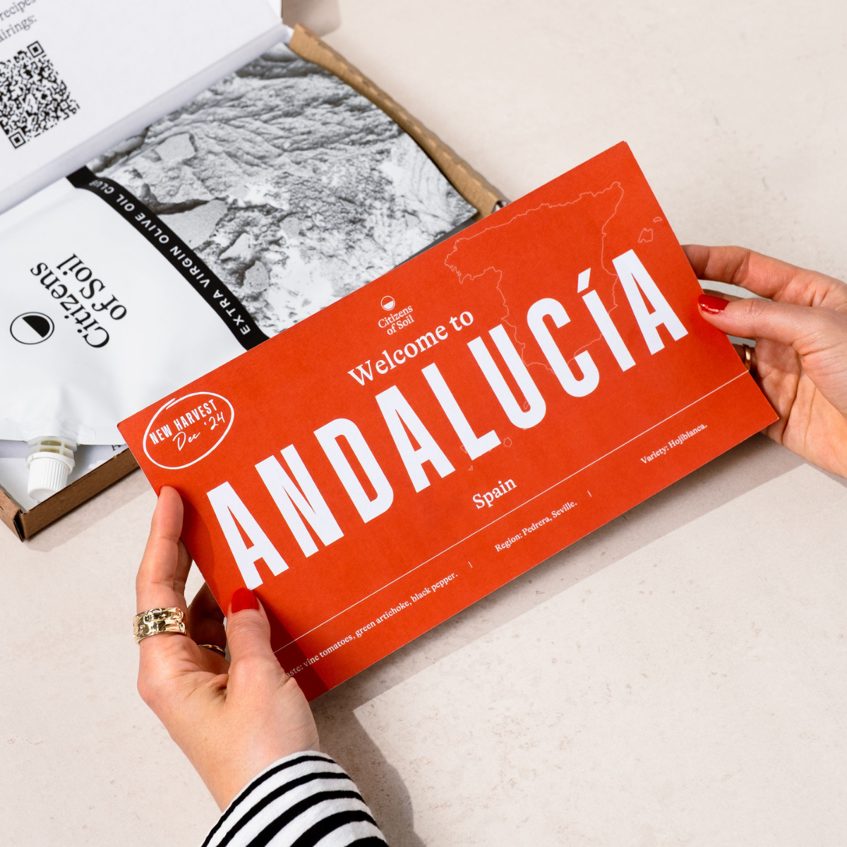 Person holding a red Olive Oil Club farmer card titled 'Welcome to Andalucóa, Spain' with a map on a light surface.