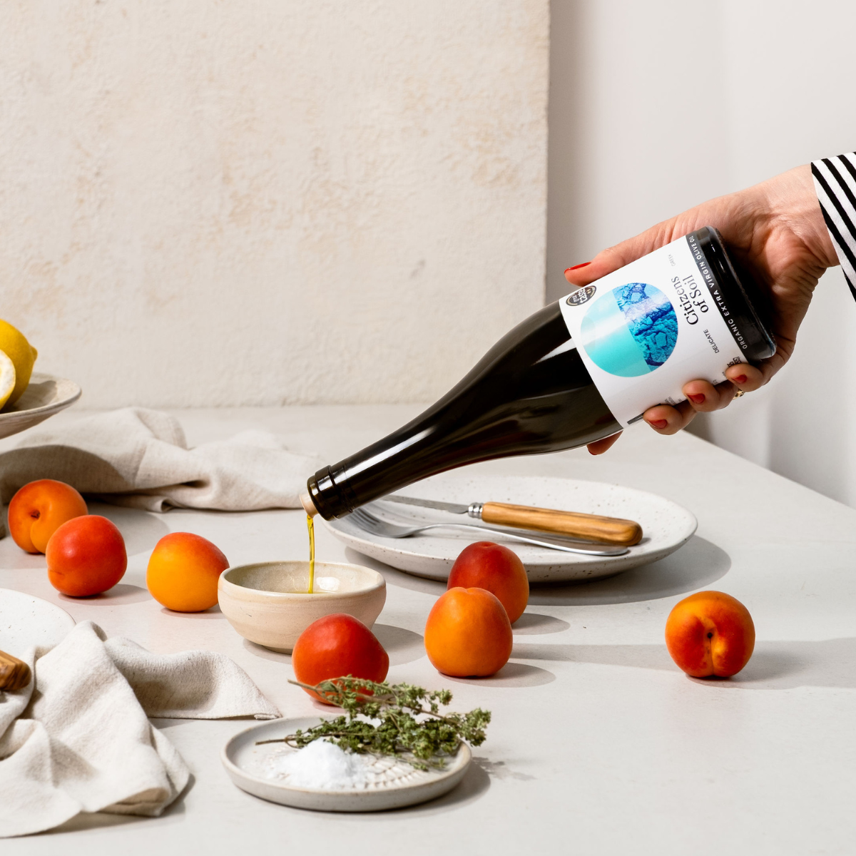 Person pouring olive oil into a glass with peaches and a bowl of herbs on a table.