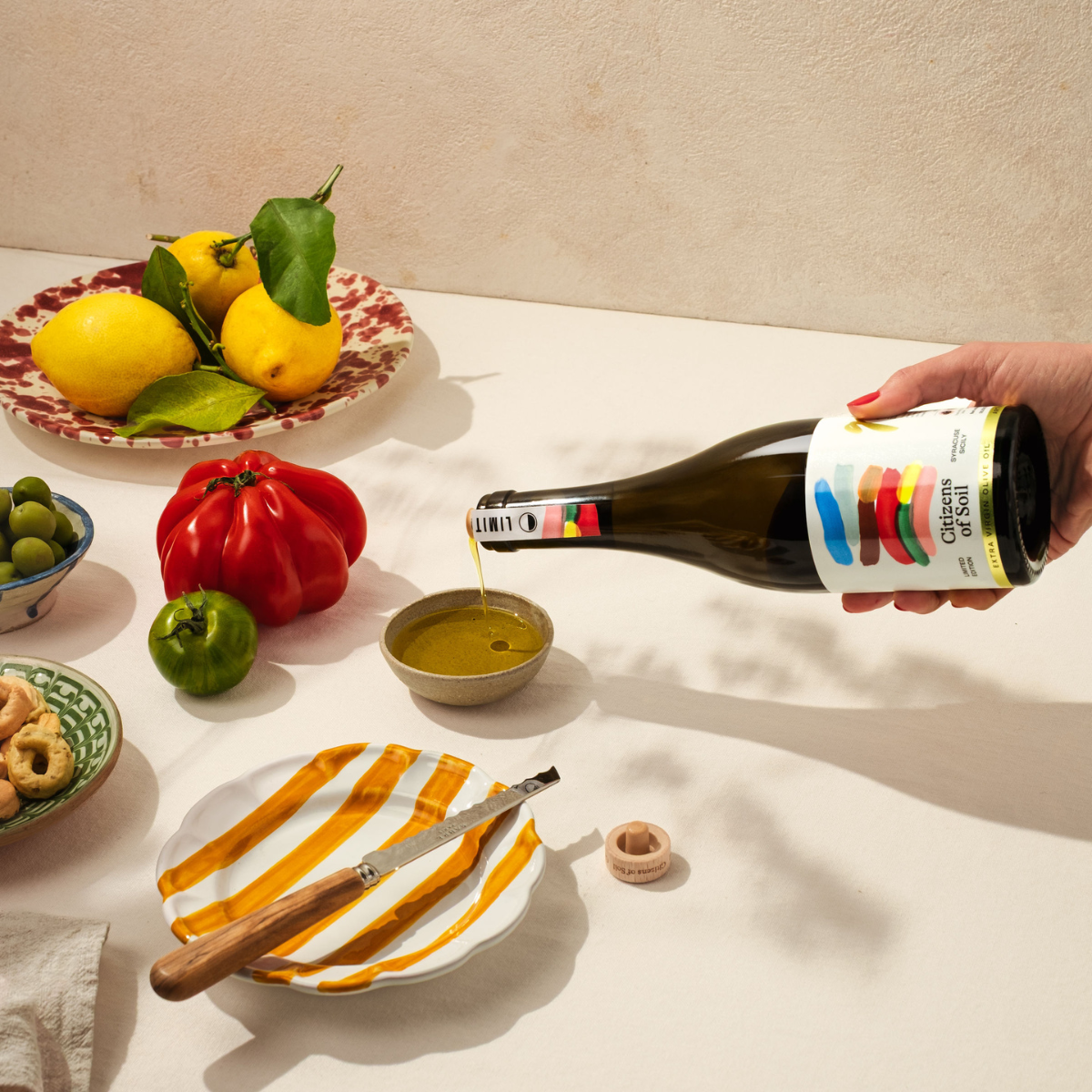Person pouring Sicilian olive oil from a bottle into a small bowl with a plate and knife on a table set with lemons, a red pepper, and other items.