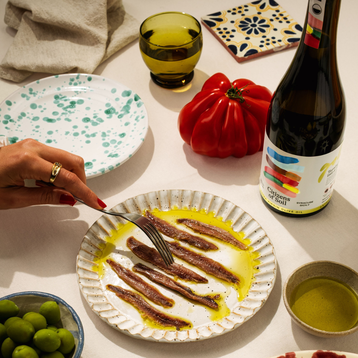 Person eating anchovies with olive oil on a plate, surrounded by a bottle of wine, tomatoes, and other items on a table.