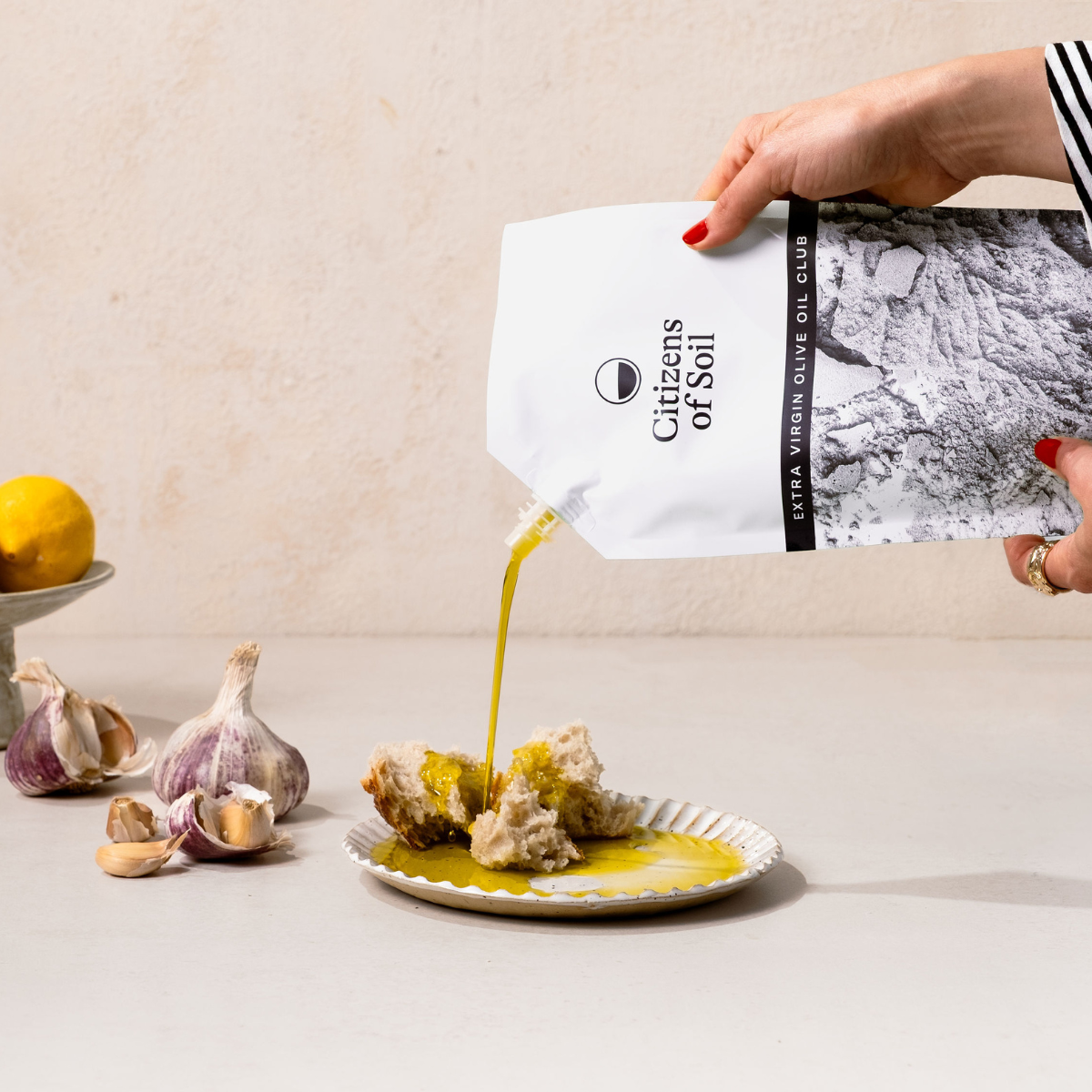 Person pouring olive oil from an Olive Oil Club pouch labeled 'Citizens of Soil' onto bread on a plate.