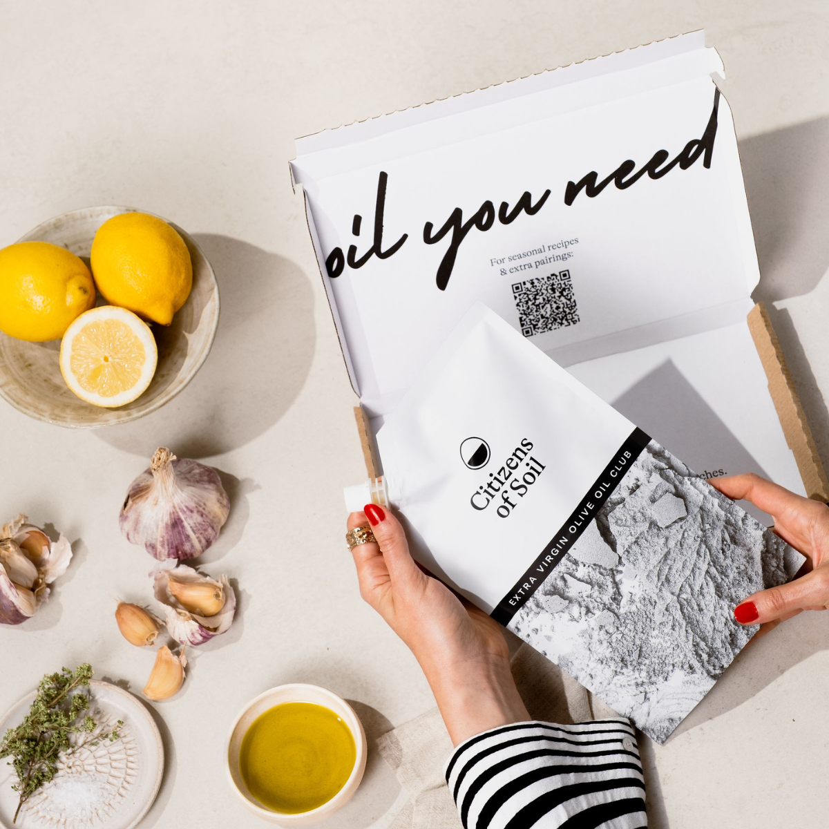 Person holding a Olive Oil Club pouch from 'Citizens of Soil' over a table with lemons and garlic.