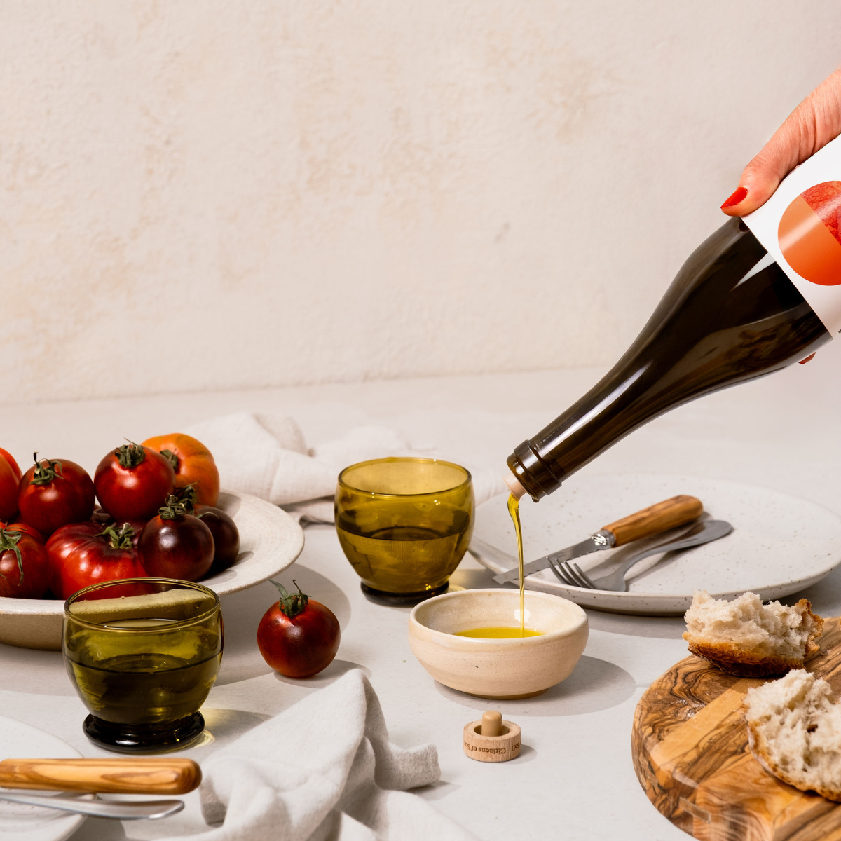 Person pouring olive oil into a small bowl with tomatoes, glasses, and bread on a table.