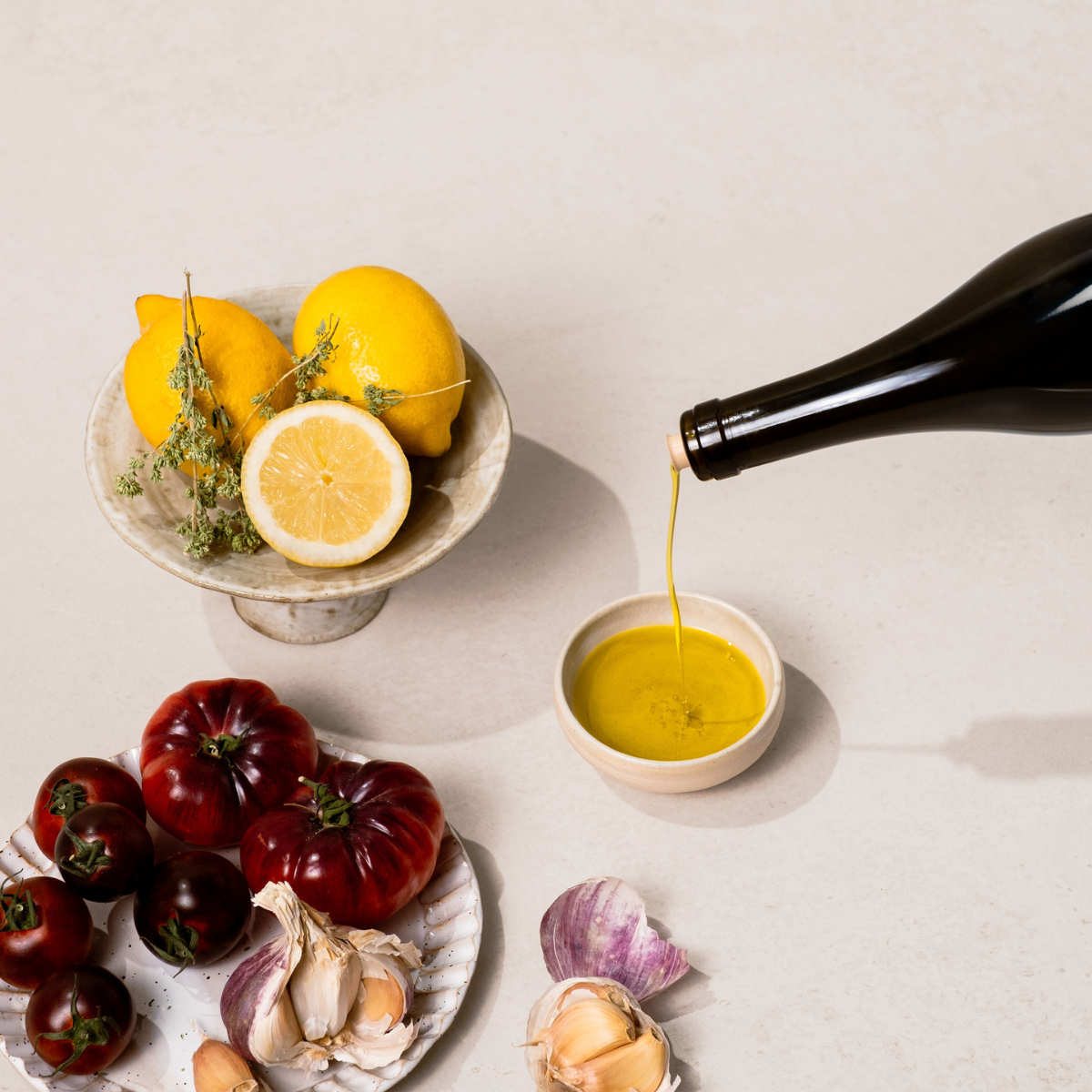 Extra virgin olive oil being poured into small bowl on white background with lemons and tomatoes on plates in the background