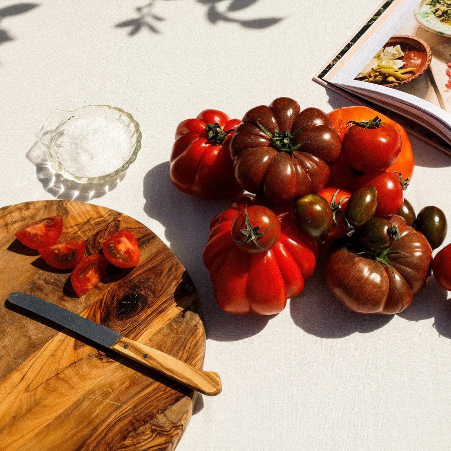 Assorted tomatoes on a wooden cutting board with a nonna knife, cookbook and olive oil container.