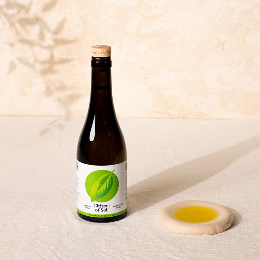 A dark glass bottle of Citizens of Soil cold-pressed oil with a cork stopper and a green basil leaf label, standing next to a small beige dish containing the yellow-green oil on a textured cream surface.