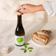 A hand with red nail polish and a gold ring is shown unscrewing a cork from a dark glass bottle labeled 'Citizens of Soil'. The bottle is set on a beige tablecloth next to a plate of bread, a small bowl of coarse salt, and fresh green basil leaves.