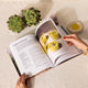An open cookbook titled 'Apple Salmorejo' on a white fabric surface, with a person's hands holding the pages. Green artichokes, a garlic bulb, a small dish of yellow oil, and the yellow prepared dishes pictured in the cookbook are arranged around it.