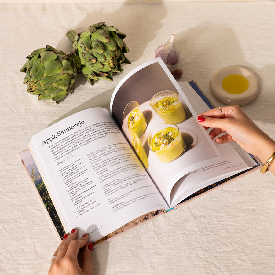 An open cookbook titled 'Apple Salmorejo' on a white fabric surface, with a person's hands holding the pages. Green artichokes, a garlic bulb, a small dish of yellow oil, and the yellow prepared dishes pictured in the cookbook are arranged around it.