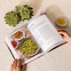 An open cookbook on a white textured fabric surface, showing a recipe page for pasta. Two hands with red nail polish hold the book open.