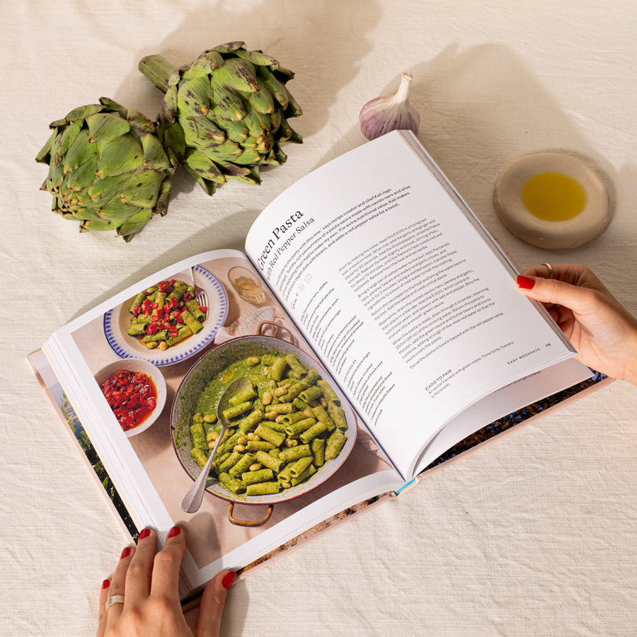 An open cookbook on a white textured fabric surface, showing a recipe page for pasta. Two hands with red nail polish hold the book open.
