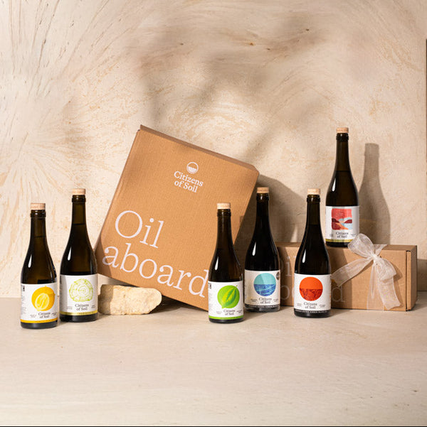 A curated gift set featuring multiple dark glass bottles of olive oil with colorful labels, a brown cardboard box labeled 'Oil aboard', and a small wrapped gift box, arranged on a light wood surface against a matching wood-paneled background.