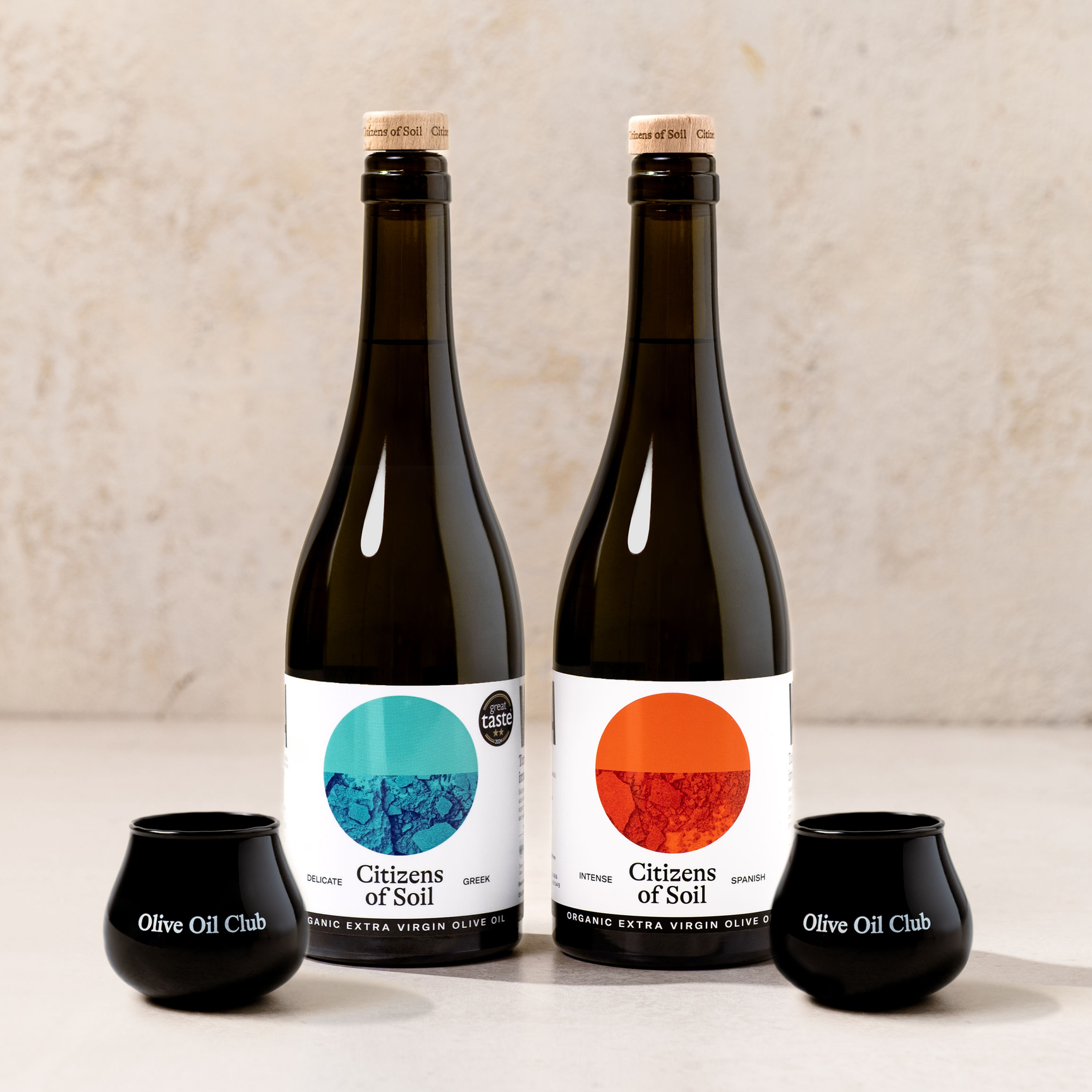Greek and Spanish bottles of 'Citizens of Soil' olive oil on a neutral background with two black Olive Oil Club tasting cups.