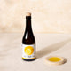 A dark glass bottle of 'Citizens of Soil' olive oil with a cork stopper and a white label featuring a yellow lemon graphic, standing next to a small beige ceramic dish filled with olive oil on a textured beige surface.