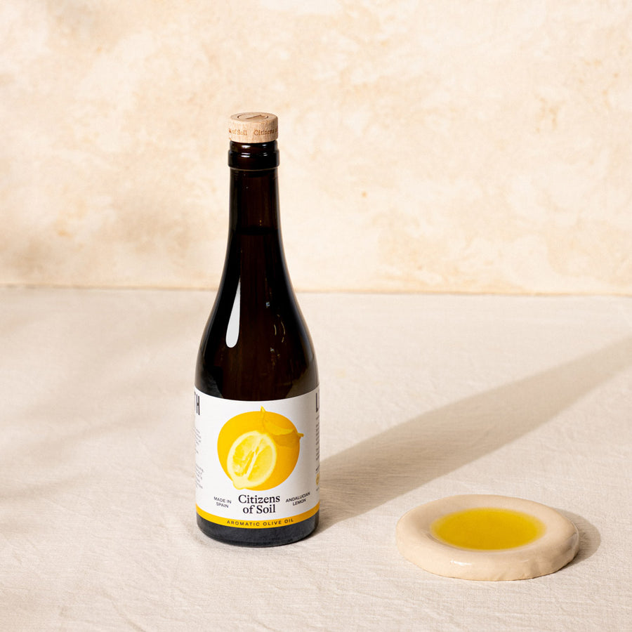 A dark glass bottle of 'Citizens of Soil' olive oil with a cork stopper and a white label featuring a yellow lemon graphic, standing next to a small beige ceramic dish filled with olive oil on a textured beige surface.