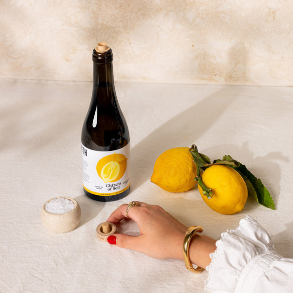 A bottle of lemon oil with a cork stopper stands on a cream fabric surface next to two yellow lemons, a small bowl of coarse salt, and a hand with red nail polish and gold jewellery opening the cork.