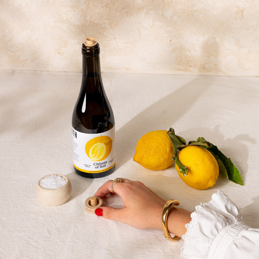 A bottle of lemon oil with a cork stopper stands on a cream fabric surface next to two yellow lemons, a small bowl of coarse salt, and a hand with red nail polish and gold jewellery opening the cork.
