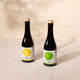 Two dark glass bottles with cork stoppers and white labels, one featuring a yellow lemon graphic and the other a green leaf graphic, set on a light beige surface against a textured beige wall with soft leaf shadows.