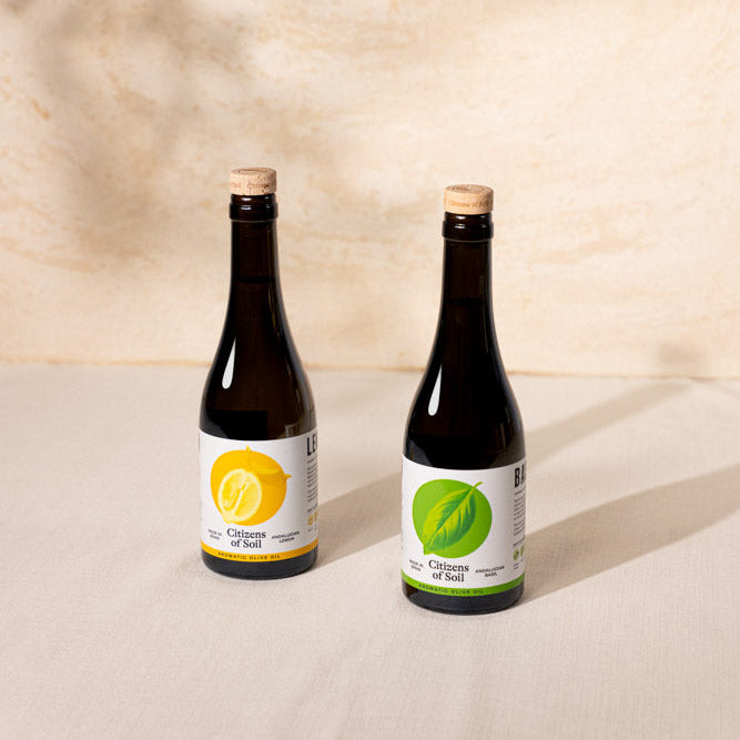 Two dark glass bottles with cork stoppers and white labels, one featuring a yellow lemon graphic and the other a green leaf graphic, set on a light beige surface against a textured beige wall with soft leaf shadows.