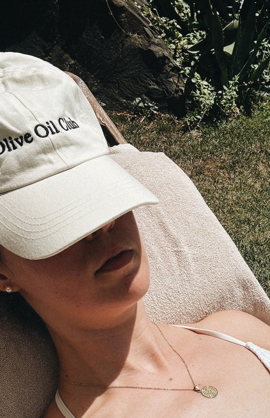 Woman laying in sun with white Olive Oil Club cap resting on face.