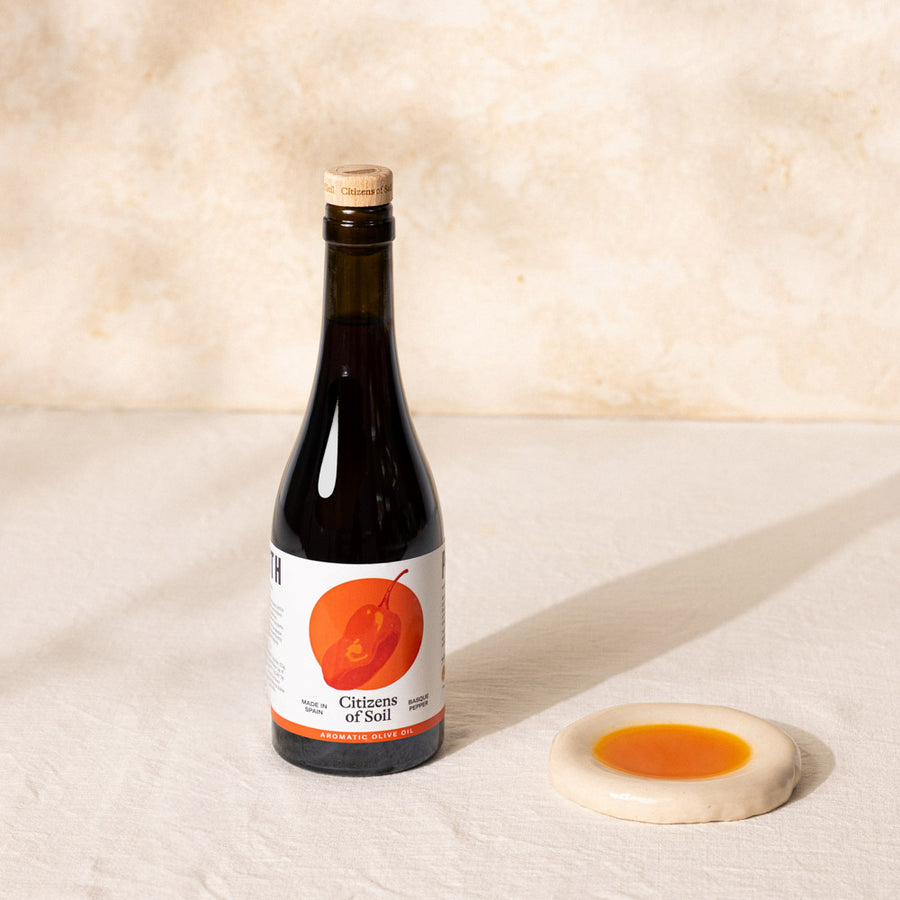 A dark glass bottle of Citizens of Soil olive oil with a cork stopper and a white label featuring an orange illustration, set on a textured beige surface next to a small ceramic dish of red pepper oil.