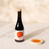 A dark glass bottle of Citizens of Soil olive oil with a cork stopper and a white label featuring an orange illustration, set on a textured beige surface next to a small ceramic dish of red pepper oil.