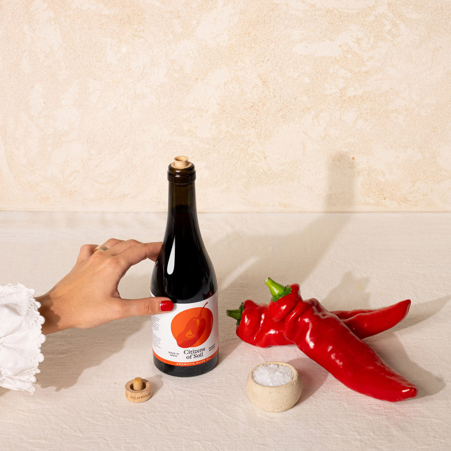 A hand with red nail polish holds a dark glass bottle labeled 'Citizens of Soil' next to two whole red chili peppers and a small bowl of salt on a textured beige surface.