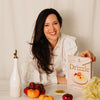 A smiling woman with long dark hair sits at a table holding a cookbook titled 'Drizzle'. She is wearing a white blouse and surrounded by fresh ingredients, including tomatoes, cabbage, and a white bottle, against a warm off-white curtain background.