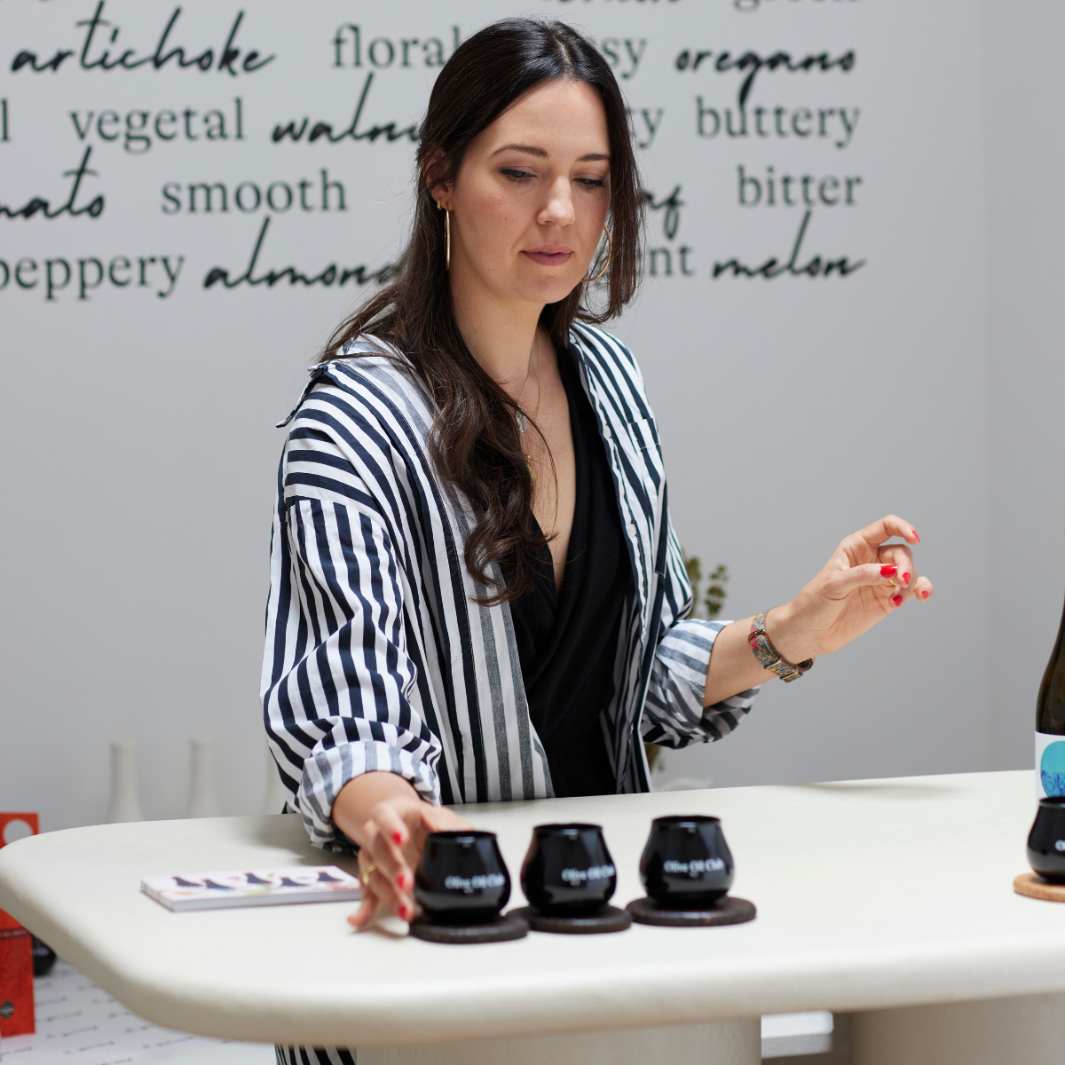 Female olive oil sommelier at a tasting table reaching towards three black tasting cups on a light clay table.