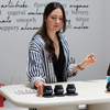 Female olive oil sommelier at a tasting table reaching towards three black tasting cups on a light clay table.