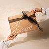 A brown cardboard gift box with the words 'Oil aboard' printed in white, tied with a sheer black ribbon, being handed from one person to another against a warm beige background.