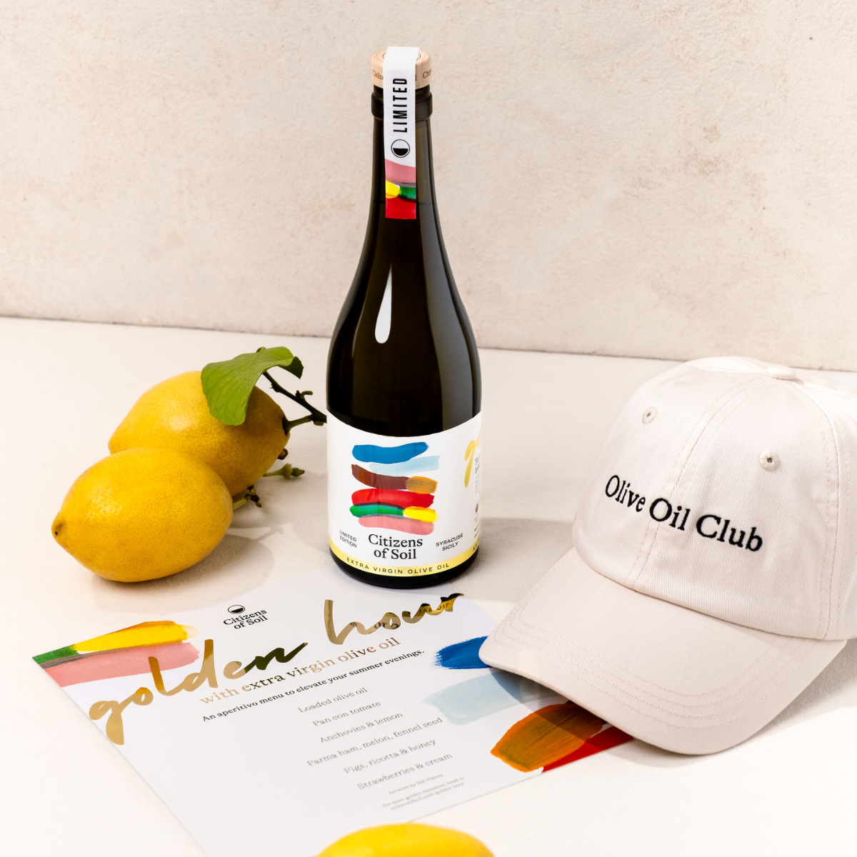 Bottle of Sicilian olive oil with a Olive Oil Club branded cap, lemons, and a magazine on a light surface.