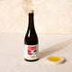 A dark glass bottle of Citizens of Soil organic olive oil with a cork stopper and a white label, displayed next to a small beige ceramic dish containing yellow liquid on a textured cream surface.