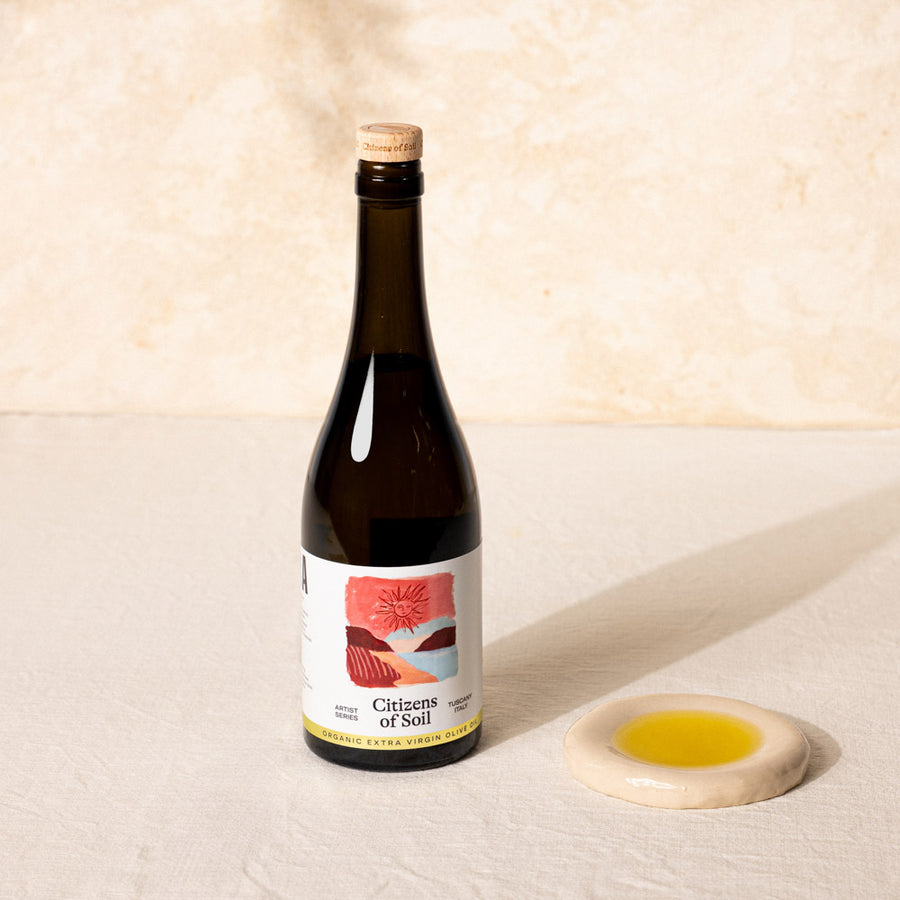 A dark glass bottle of Citizens of Soil organic olive oil with a cork stopper and a white label, displayed next to a small beige ceramic dish containing yellow liquid on a textured cream surface.