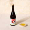 A dark glass bottle of Citizens of Soil organic olive oil with a cork stopper and a white label, displayed next to a small beige ceramic dish containing yellow liquid on a textured cream surface.