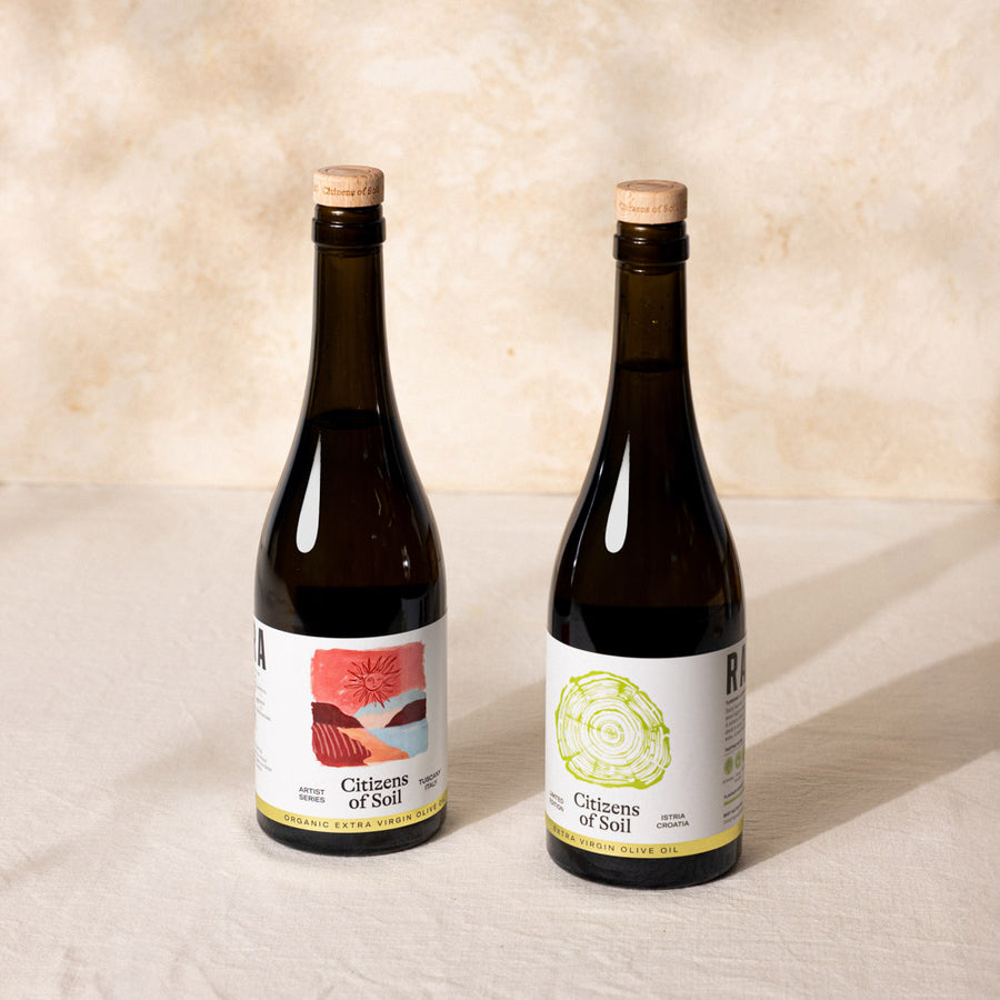 Two dark glass bottles of Citizens of Soil organic olive oil with cork stoppers, standing side-by-side on a white fabric surface against a textured beige background.