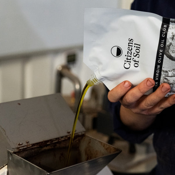 Person pouring olive oil from a 'Citizens of Soil' bag into a chocolate machine.