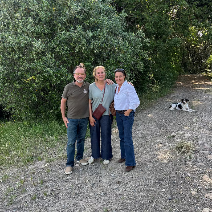 Three people posing together outdoors on a gravel path surrounded by green bushes, with a black and white dog lying in the background.