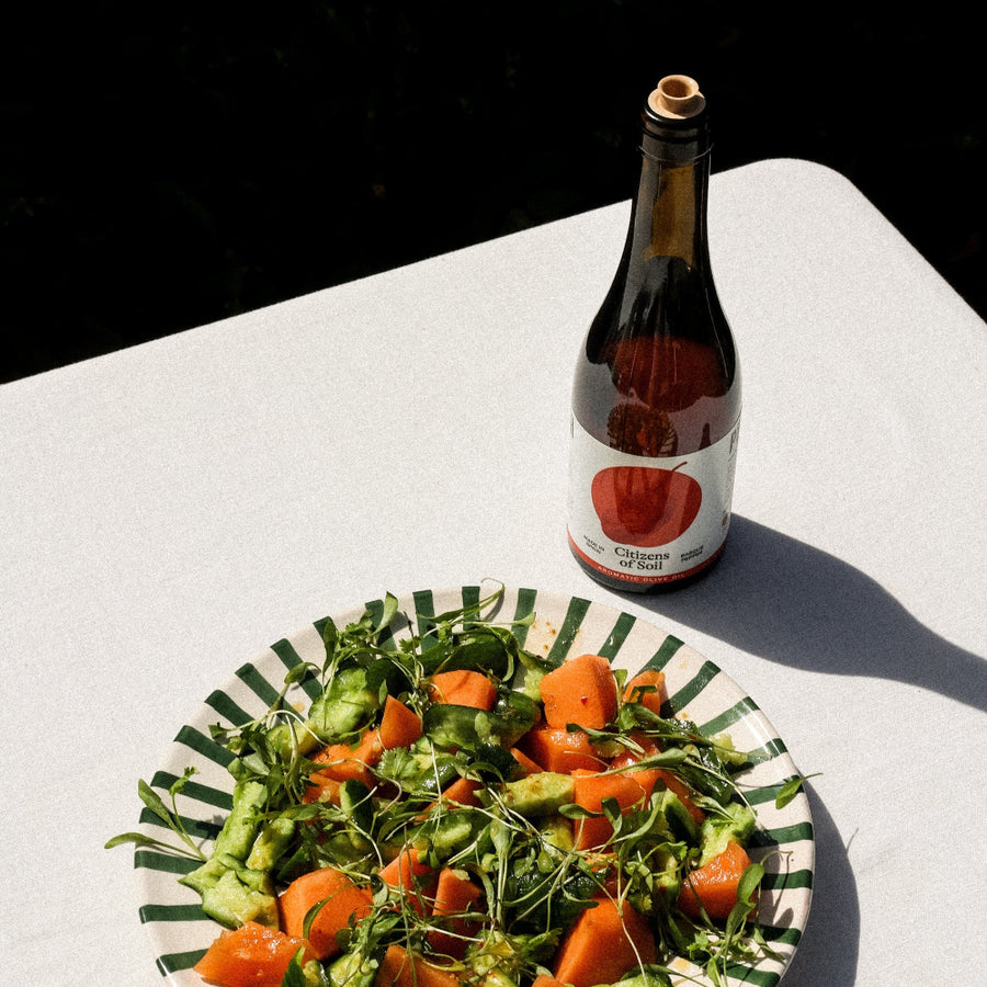 A bottle of olive oil with a cork stopper and a striped paper plate of mixed green and orange salad on a white tablecloth, photographed in bright natural sunlight.