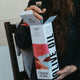 A person holds an open box of Citizens of Soil organic extra virgin olive oil from Tuscany, Italy. The packaging features a white background with a red and blue abstract floral design and bold black text reading 'OLIVE OIL'.
