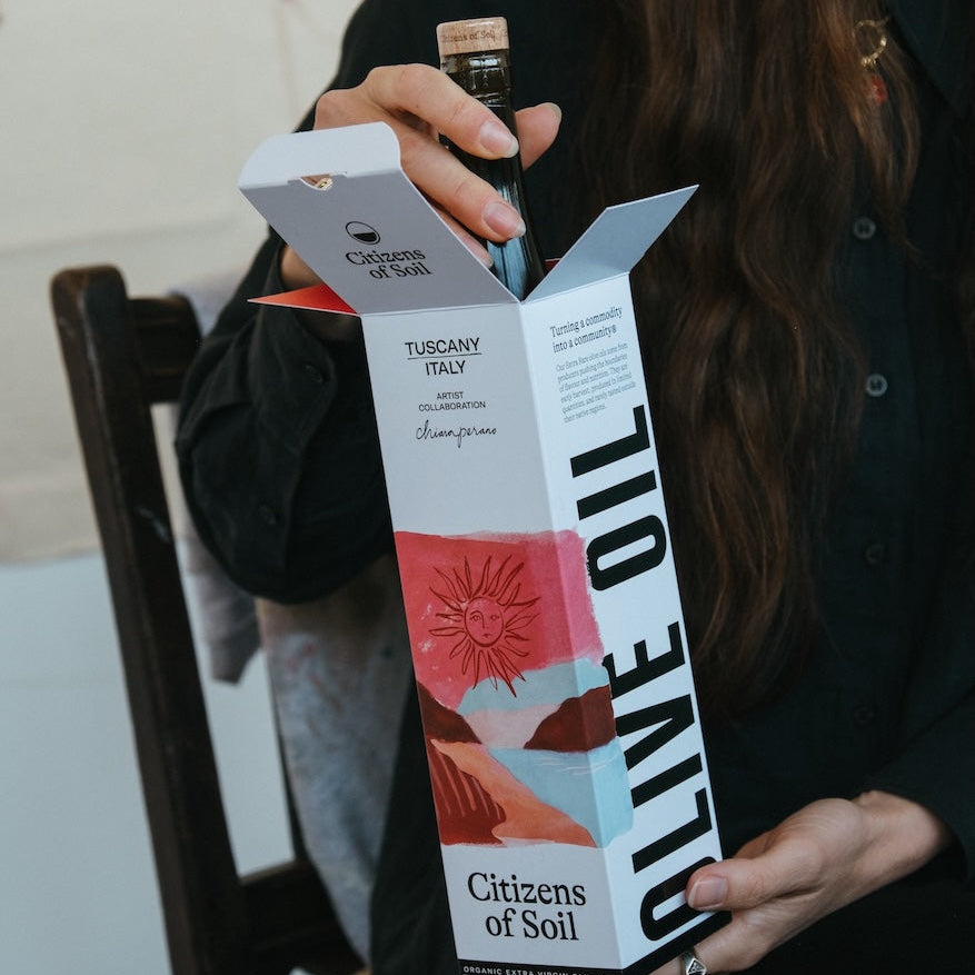 A person holds an open box of Citizens of Soil organic extra virgin olive oil from Tuscany, Italy. The packaging features a white background with a red and blue abstract floral design and bold black text reading 'OLIVE OIL'.